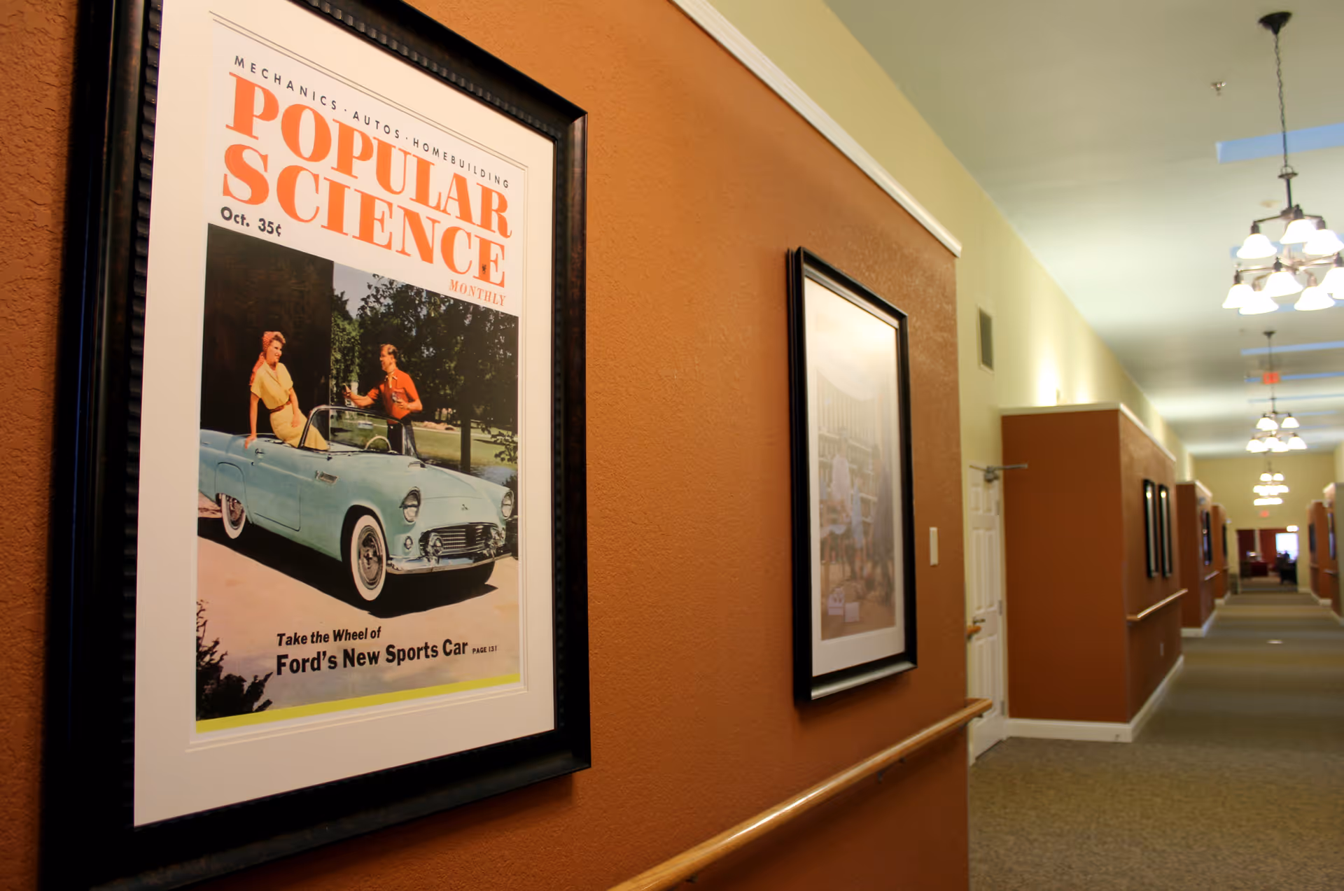 A long hallway in a senior living facility with brown and beige walls, framed vintage posters on the walls, handrails, and ceiling lights. The hallway extends into the distance with doors and more framed pictures along the walls.