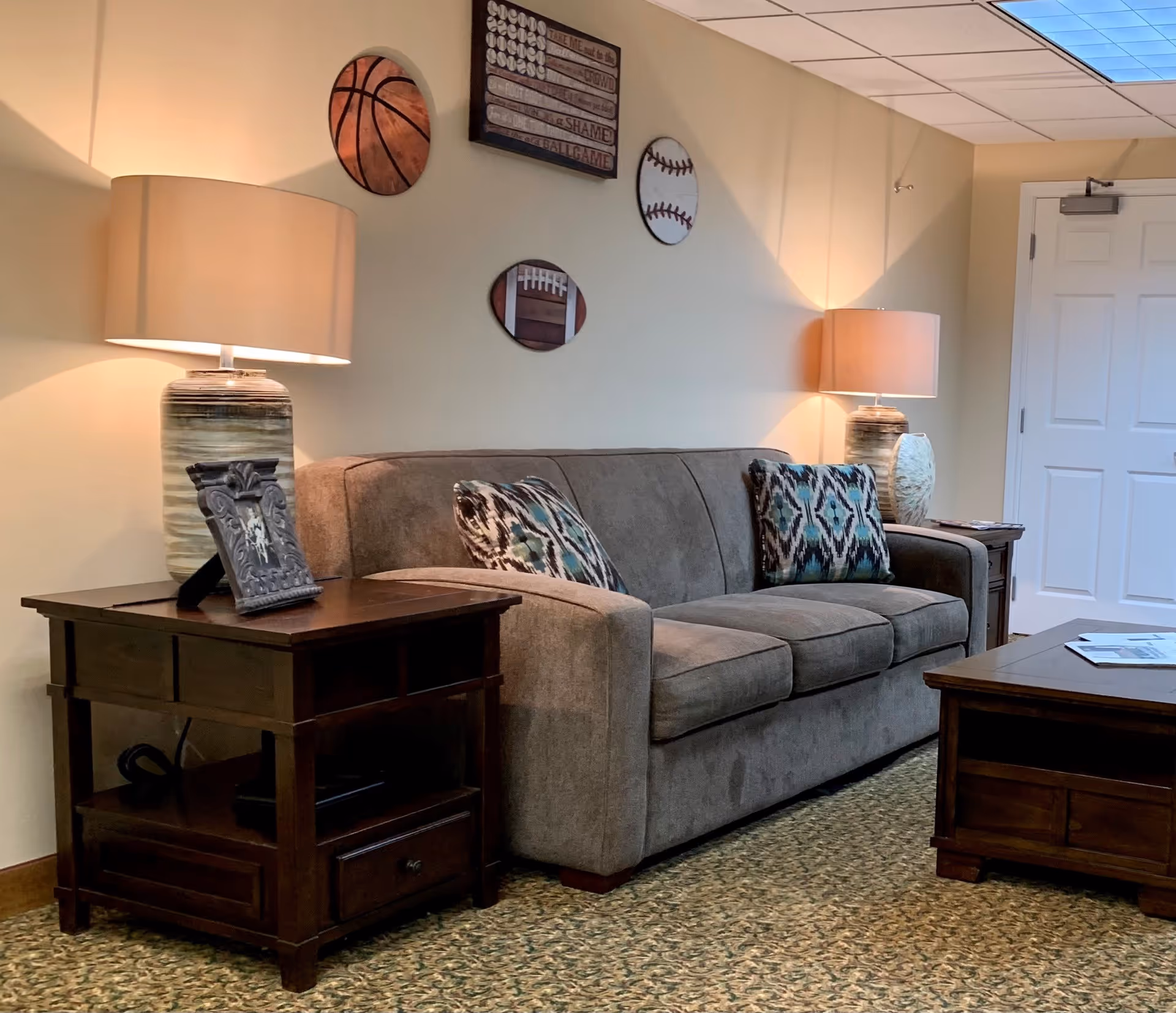 A cozy living room area with a gray sofa adorned with patterned cushions, two wooden side tables each holding a lamp, and a wooden coffee table. The wall behind the sofa features sports-themed decorations including a basketball, baseball, football, and a wooden plaque with text. The room has a carpeted floor and a white door in the background.