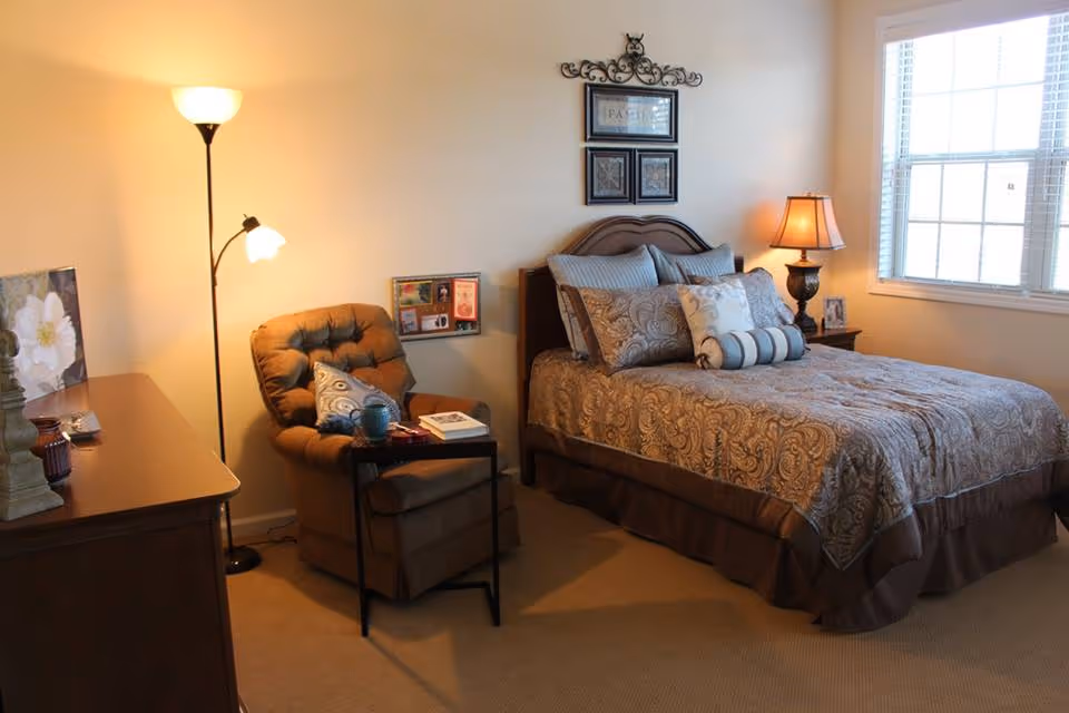 A cozy bedroom with a neatly made bed featuring patterned bedding and multiple pillows. Next to the bed is a wooden nightstand with a lamp and a framed photo. A comfortable brown armchair with a small side table holding a cup and a book is positioned near a tall floor lamp. The room has beige walls and carpet, and a window with white blinds lets in natural light.