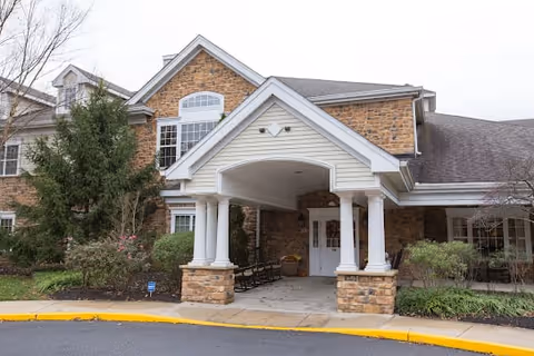 Front exterior view of a senior living facility named Spring Home at Galloway, featuring a covered entrance with white columns, stone and siding facade, and surrounding greenery.