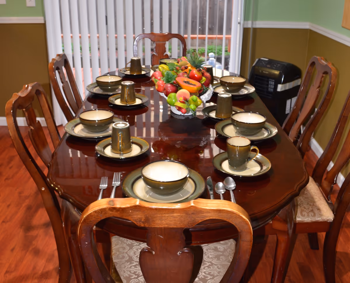 A polished wooden dining table set for eight with matching wooden chairs. Each place setting includes a plate, bowl, cup, and utensils. A decorative centerpiece with artificial fruits is placed in the middle of the table. Vertical blinds cover a window in the background, and a portable heater is positioned against the wall.