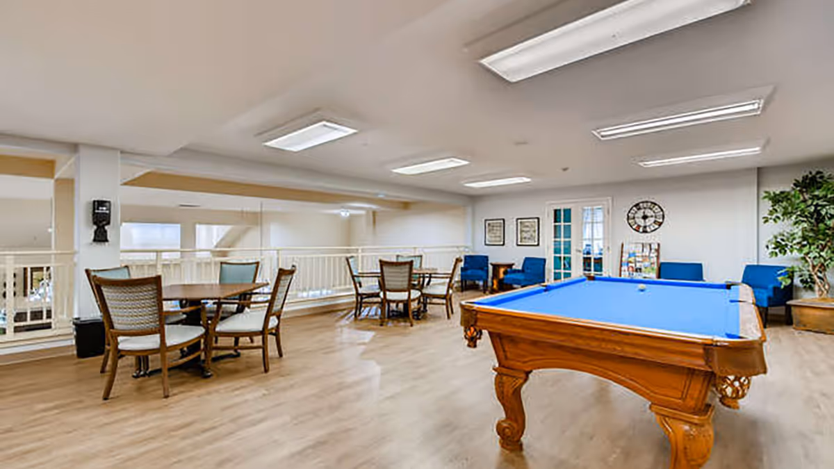 A spacious recreational room with a blue felt pool table in the foreground, several round tables with chairs, blue armchairs along the back wall, framed pictures, a large clock, and a potted plant in the corner. The room has wood flooring and bright overhead lighting.