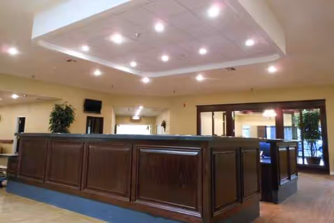 Interior lobby with a dark wooden reception desk, bright ceiling lights, and potted plants.