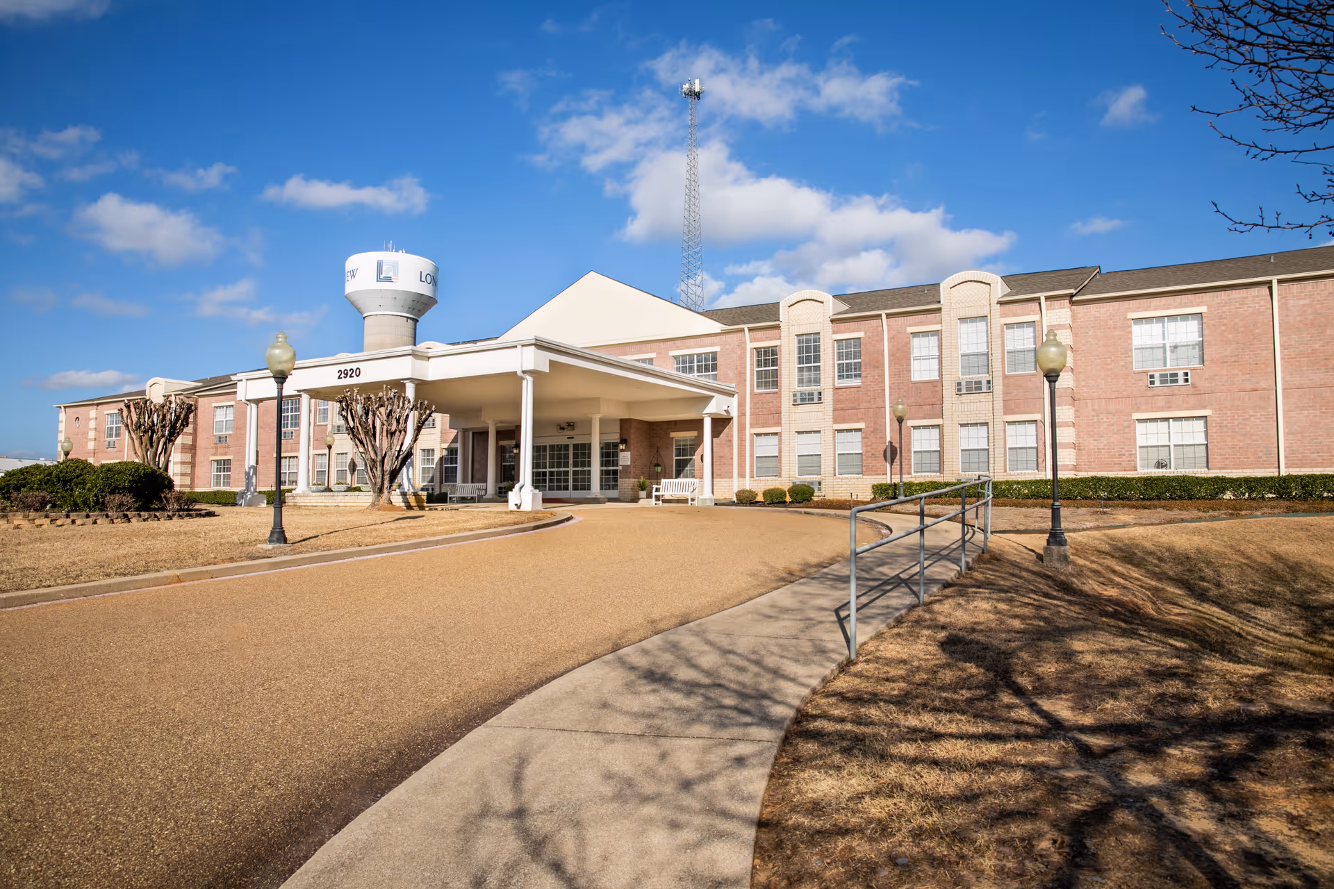 Exterior view of a two-story brick senior living facility building with a covered entrance, a paved driveway, lamp posts, trimmed bushes, and a water tower in the background under a blue sky with some clouds.