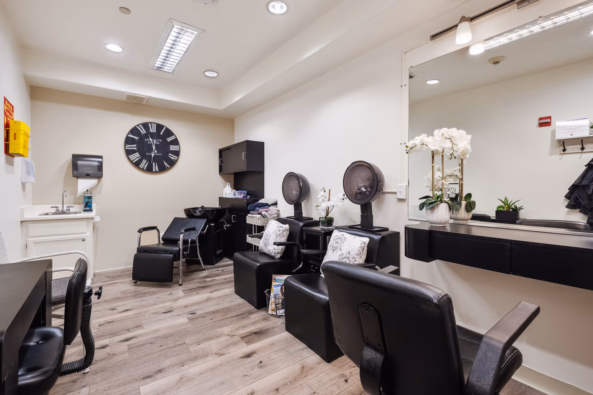 Interior of a salon room with black salon chairs, hair dryers, a large wall mirror, a sink, and a wall clock. The room has light-colored walls and wood flooring, with some decorative plants and pillows on the chairs.