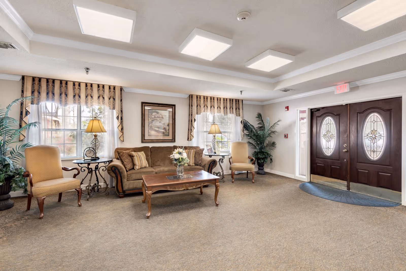 A well-lit living room area with a brown sofa, two beige armchairs, a wooden coffee table with a flower vase, two side tables with lamps, large windows with patterned curtains, potted plants, and double wooden doors with decorative glass panels.