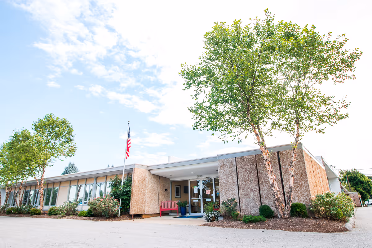 Exterior view of a single-story building with large windows, an American flag on a flagpole, and several trees and shrubs around the entrance. There is a red bench near the entrance door under a covered walkway.