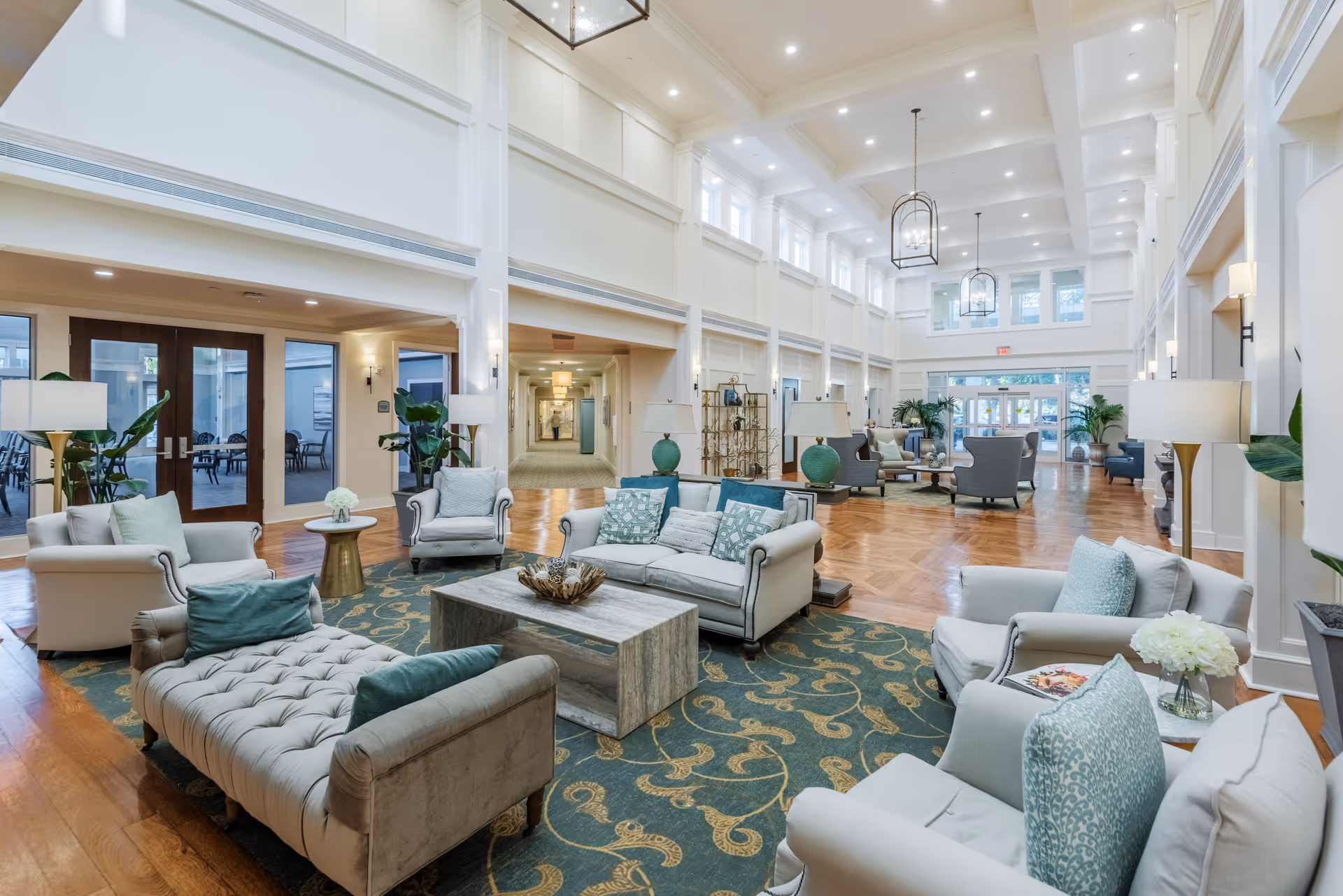Spacious and elegant living room area in a senior living facility with high ceilings, large windows, and multiple seating arrangements including sofas, armchairs, and a cushioned bench. The room features wooden flooring, decorative rugs, lamps, and plants, creating a welcoming and comfortable atmosphere.
