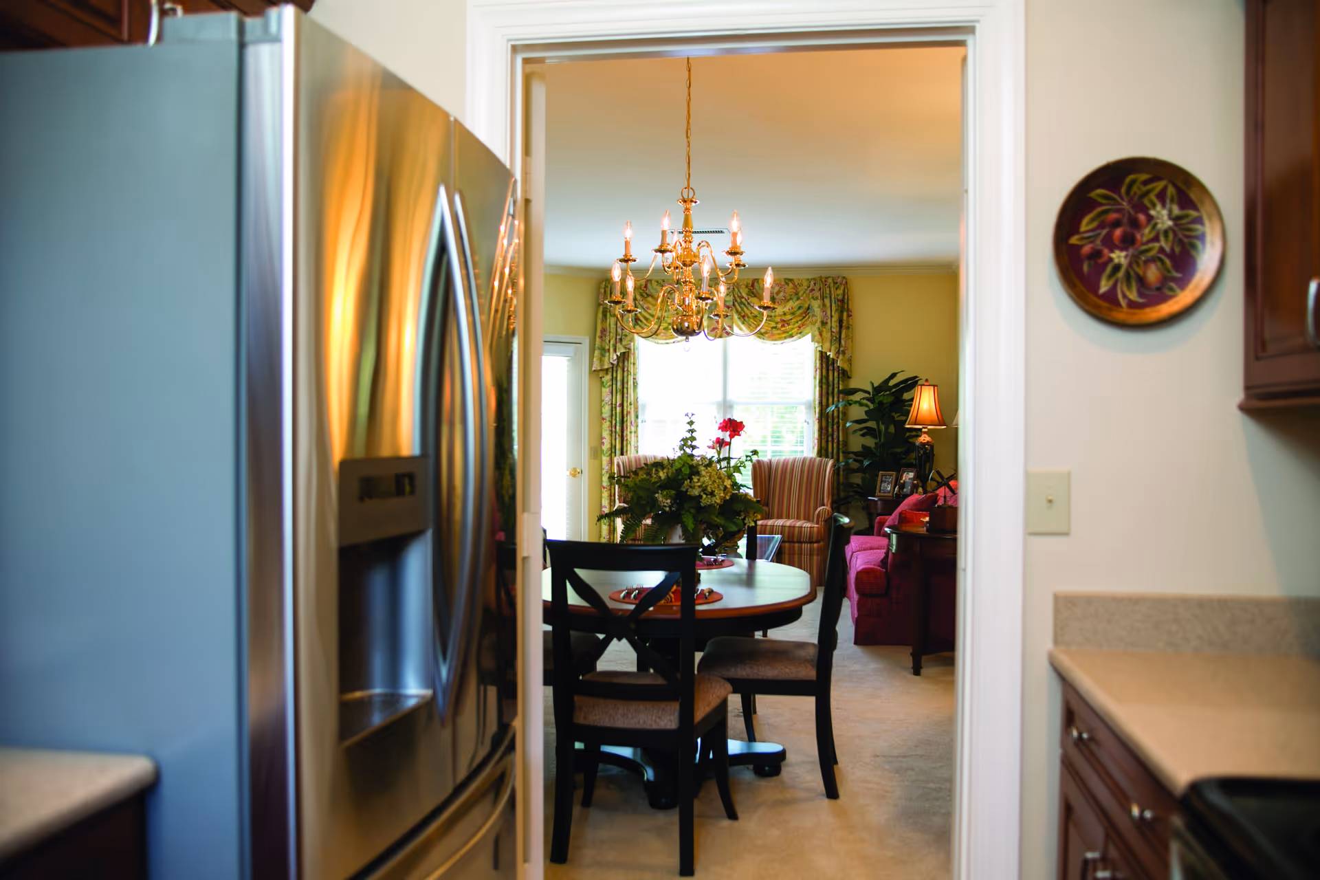 View from a kitchen into a dining and living area. The kitchen features a stainless steel refrigerator and wooden cabinets. The dining area has a round table with four chairs and a floral centerpiece. A chandelier hangs above the table. The living area has a striped armchair, a red sofa, a side table with a lamp, and a large window with floral curtains.