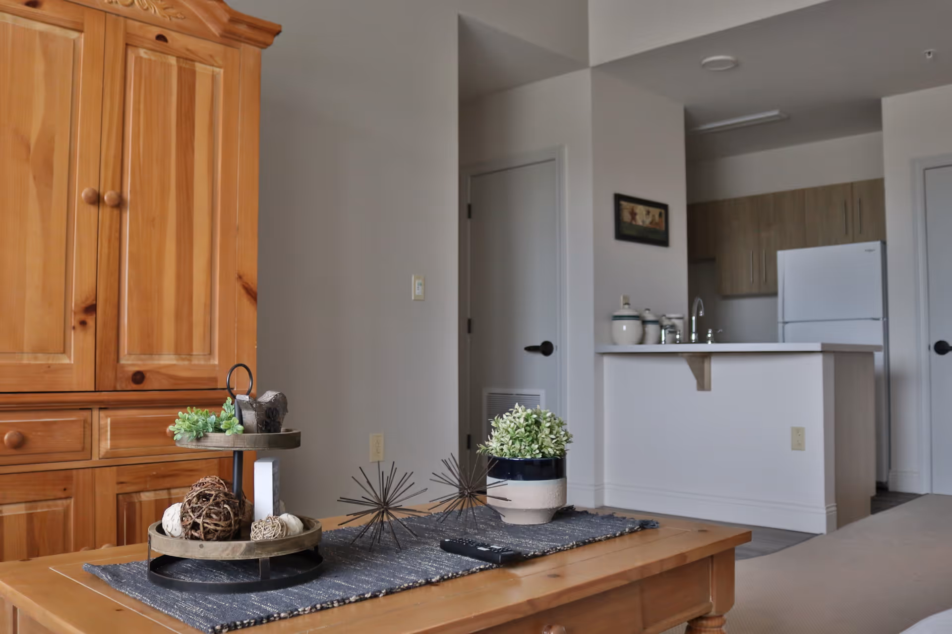 Interior view of a senior living facility unit showing a wooden coffee table with decorative items including a small plant and ornamental spheres. In the background, there is a kitchen area with a refrigerator, cabinets, and a sink, as well as a wooden cabinet on the left side.