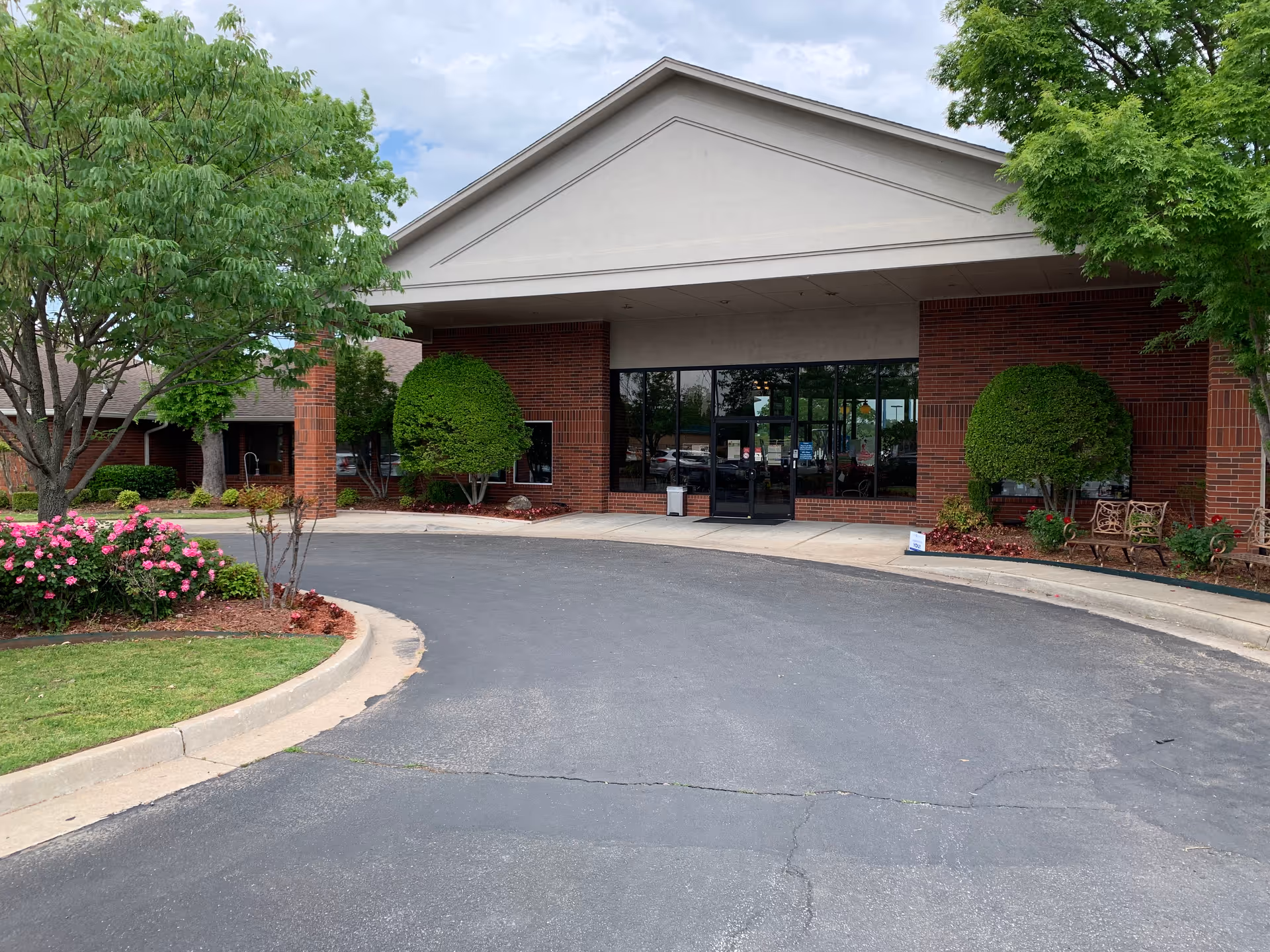 Front entrance of a brick senior living facility with a covered porte-cochere, glass doors, trimmed shrubs and a circular driveway.