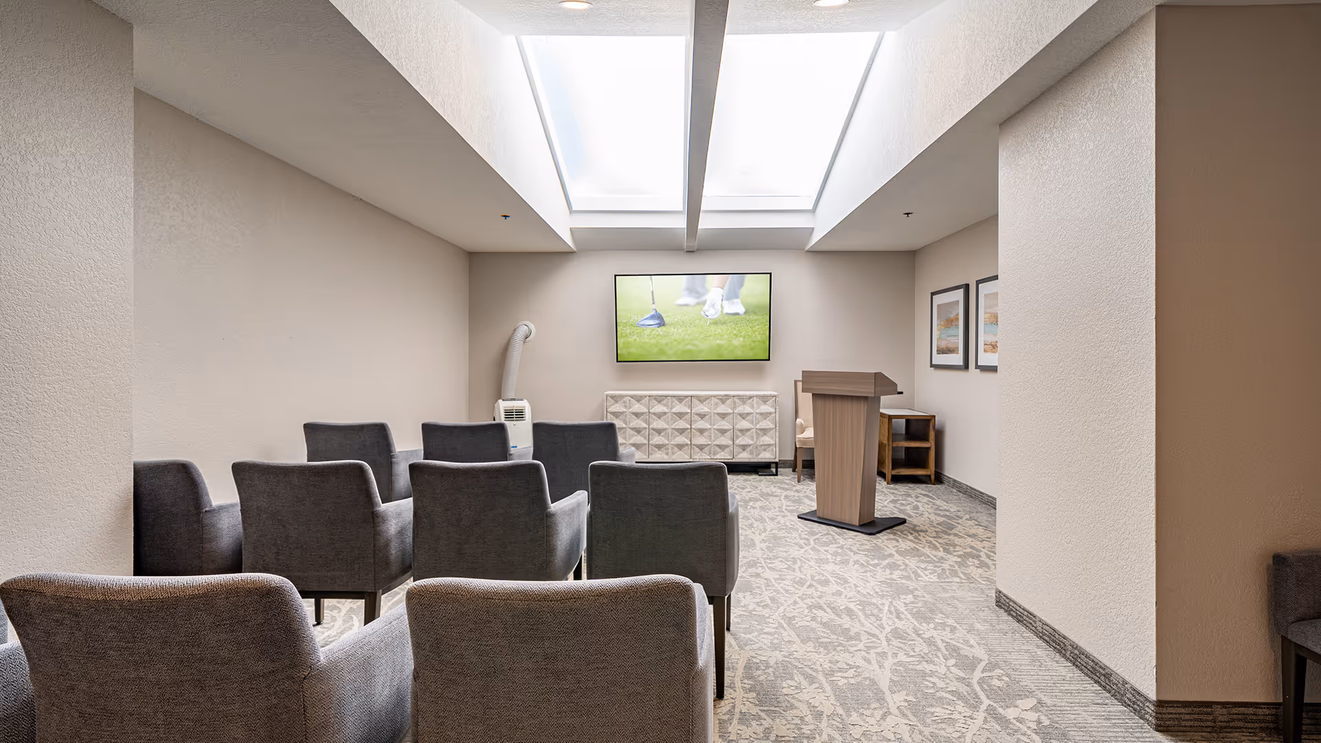 A small meeting or presentation room with two rows of gray upholstered chairs facing a wooden podium and a wall-mounted flat screen TV displaying a golf scene. The room has beige walls, carpeted floor with a subtle pattern, two skylights on the ceiling, and framed artwork on the right wall.