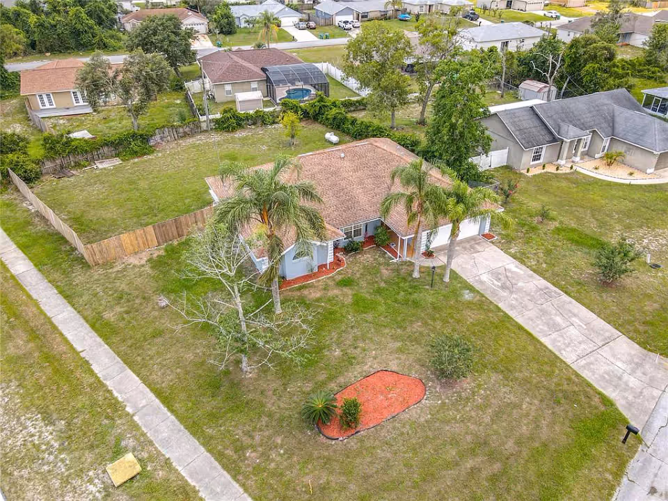 Aerial view of a single-story house with a brown roof, surrounded by a grassy yard with palm trees and a small landscaped area with red mulch. The house has a driveway leading to a garage and is located in a suburban neighborhood with other houses nearby.