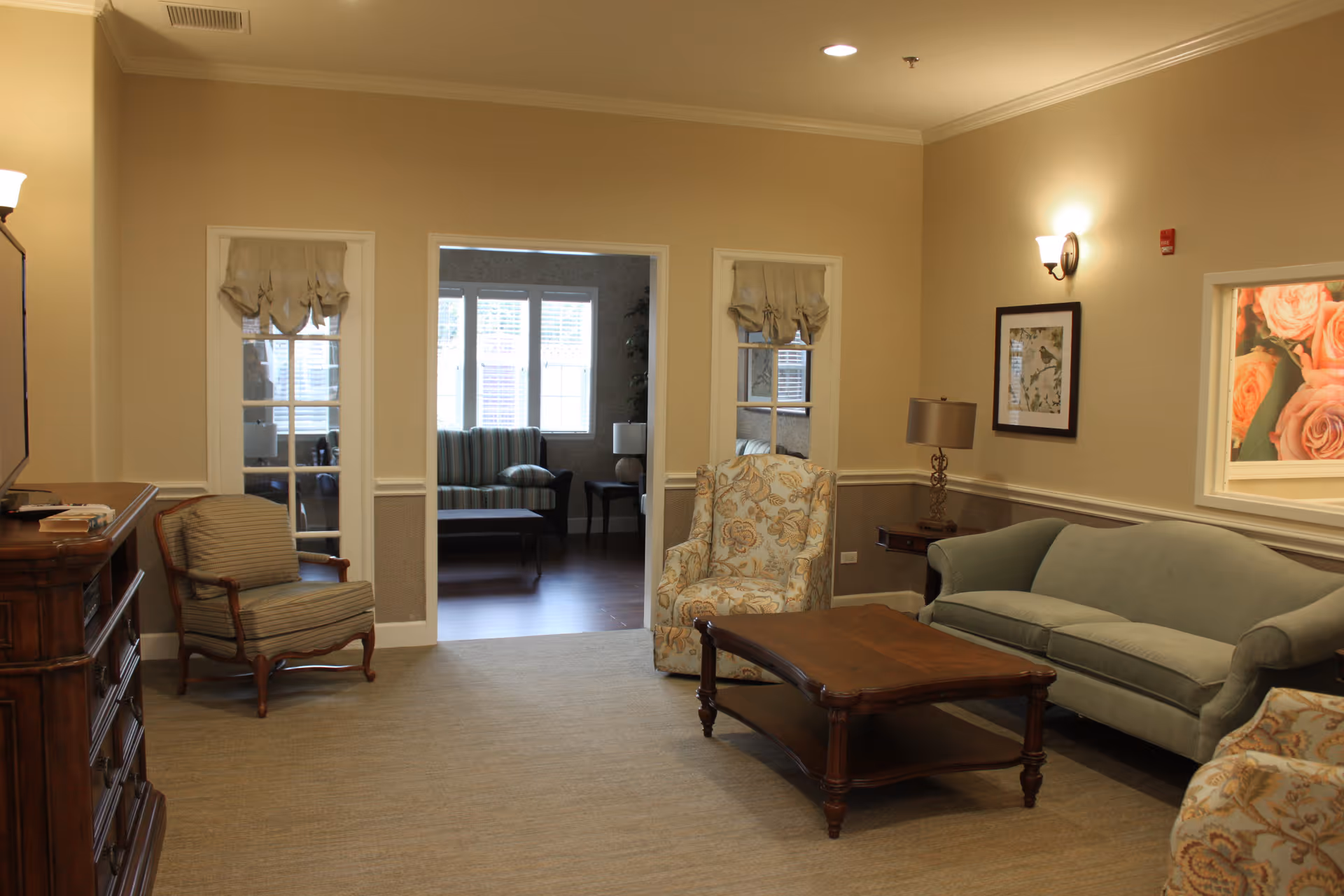 A cozy living room area in a senior living facility with beige walls and carpeted floor. The room features a green upholstered sofa, two patterned armchairs, a wooden coffee table, a wooden side table with a lamp, framed artwork on the walls, and a doorway leading to another room with additional seating and windows letting in natural light.