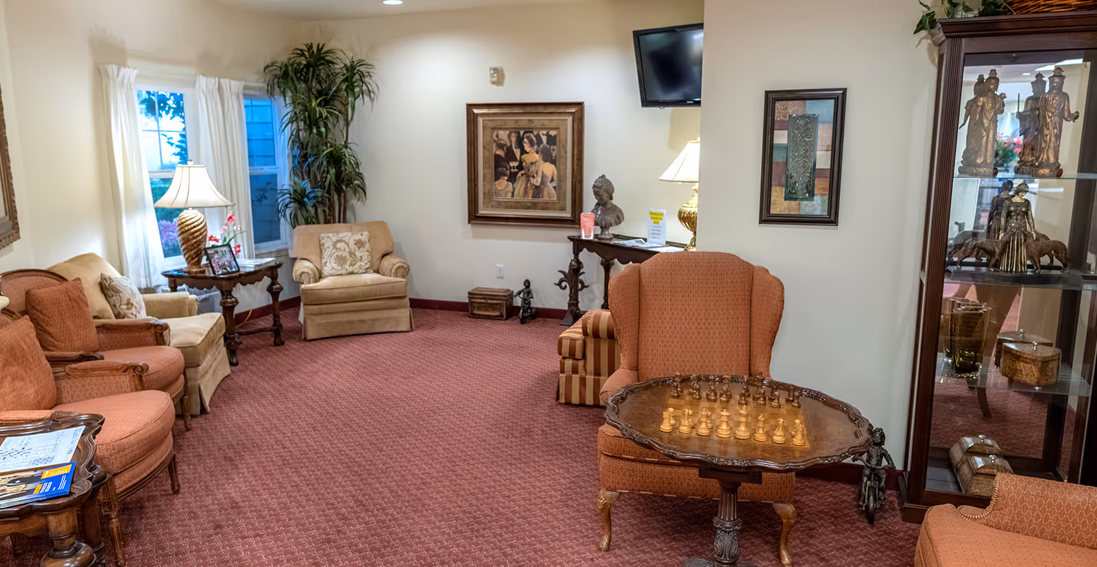 A cozy living room area with multiple upholstered armchairs and sofas arranged around small wooden tables. One table has a chess set on it. The room has red carpet flooring, cream-colored walls, framed artwork, a tall potted plant, a lamp on a side table, and a glass display cabinet with decorative statues. A flat-screen TV is mounted on the wall.
