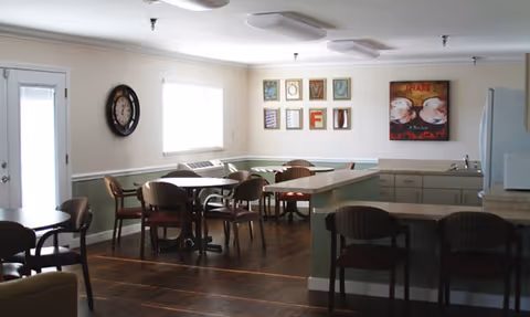 A well-lit dining area with several tables and chairs arranged on a wooden floor. The walls are painted light green with white trim, and there are framed artworks and a clock hanging on the walls. A counter with bar stools separates the dining space from a kitchenette area with cabinets and a refrigerator.
