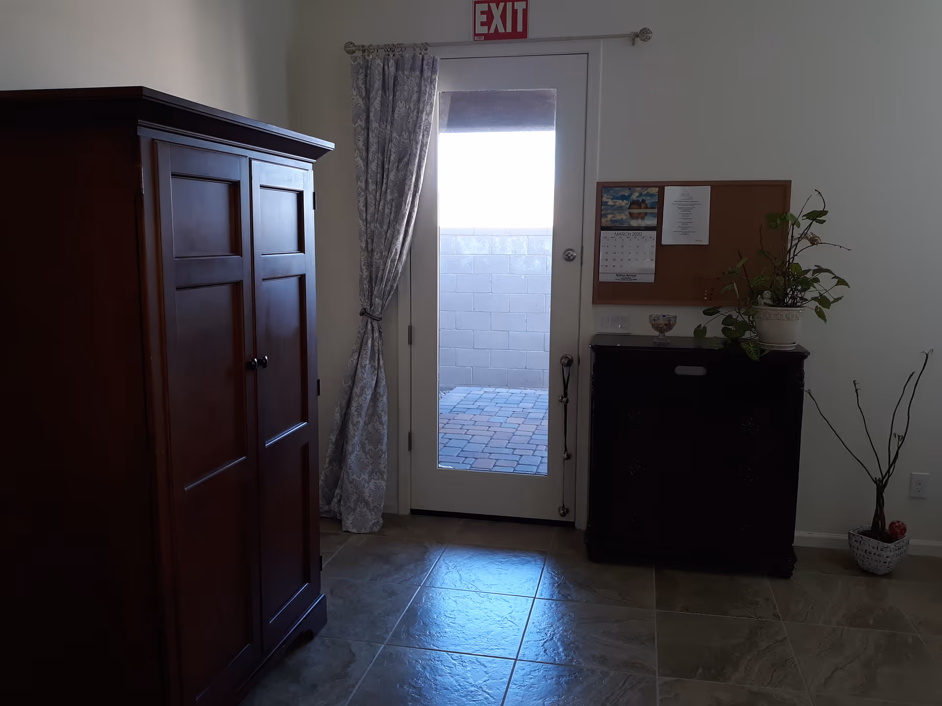 Interior view of a room with a glass door leading outside. The door has a curtain tied to one side and an exit sign above it. To the left of the door is a large wooden cabinet, and to the right is a smaller dark wooden dresser with a plant and a bulletin board above it. There is also a decorative plant in a basket on the floor to the right.