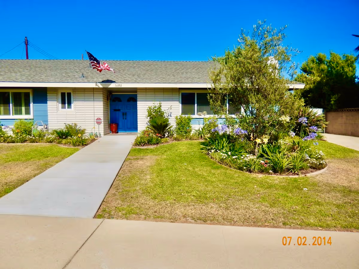 Front exterior view of a single-story residential building with a blue door, an American flag mounted near the roof, and a well-maintained lawn with a circular flower bed containing various plants and flowers.
