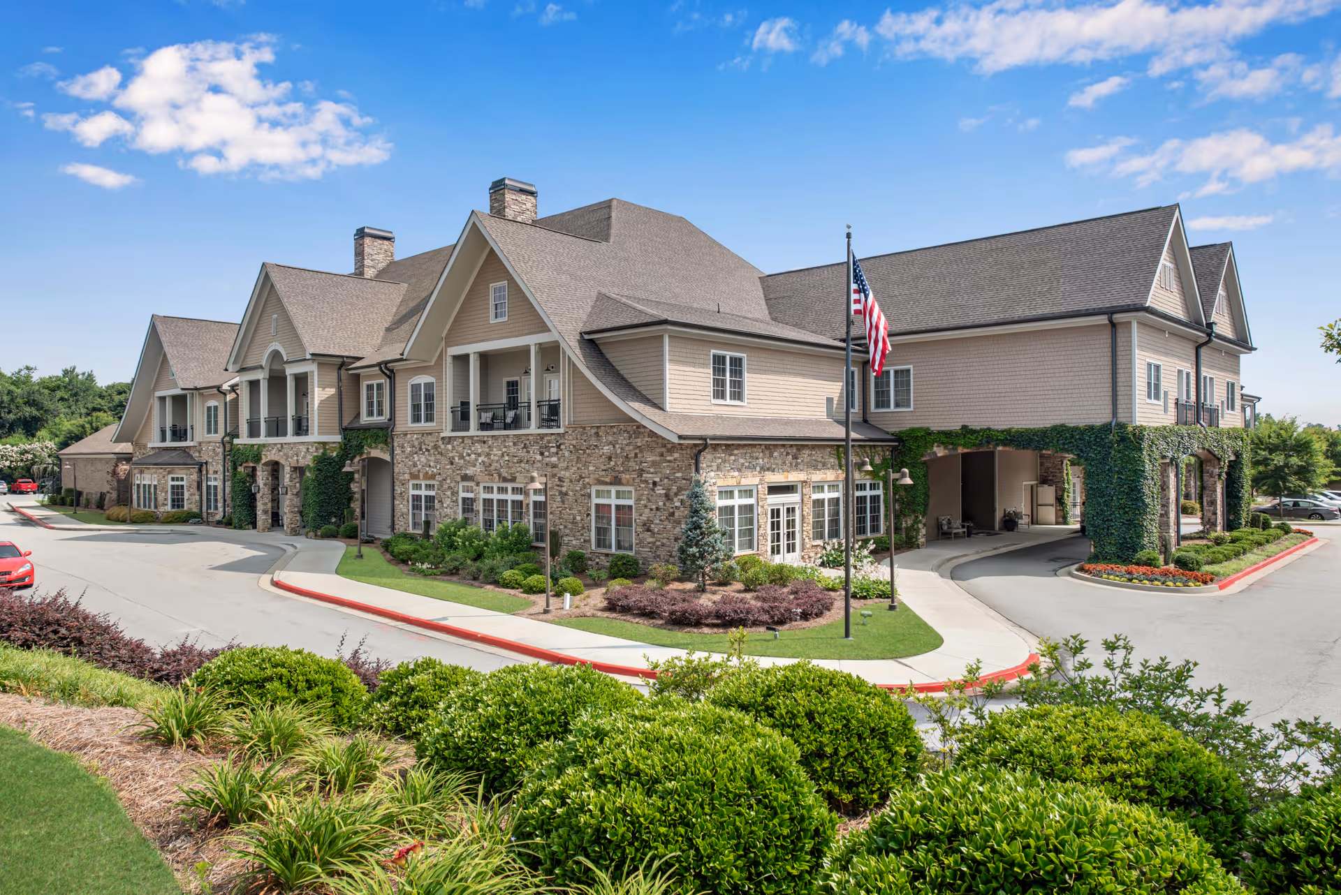 Exterior view of Dogwood Forest Grayson, a large senior living facility building with stone and beige siding, multiple windows, and a covered entrance. The building is surrounded by well-maintained landscaping including bushes, flowers, and a green lawn. An American flag is displayed on a flagpole near the entrance under a clear blue sky.