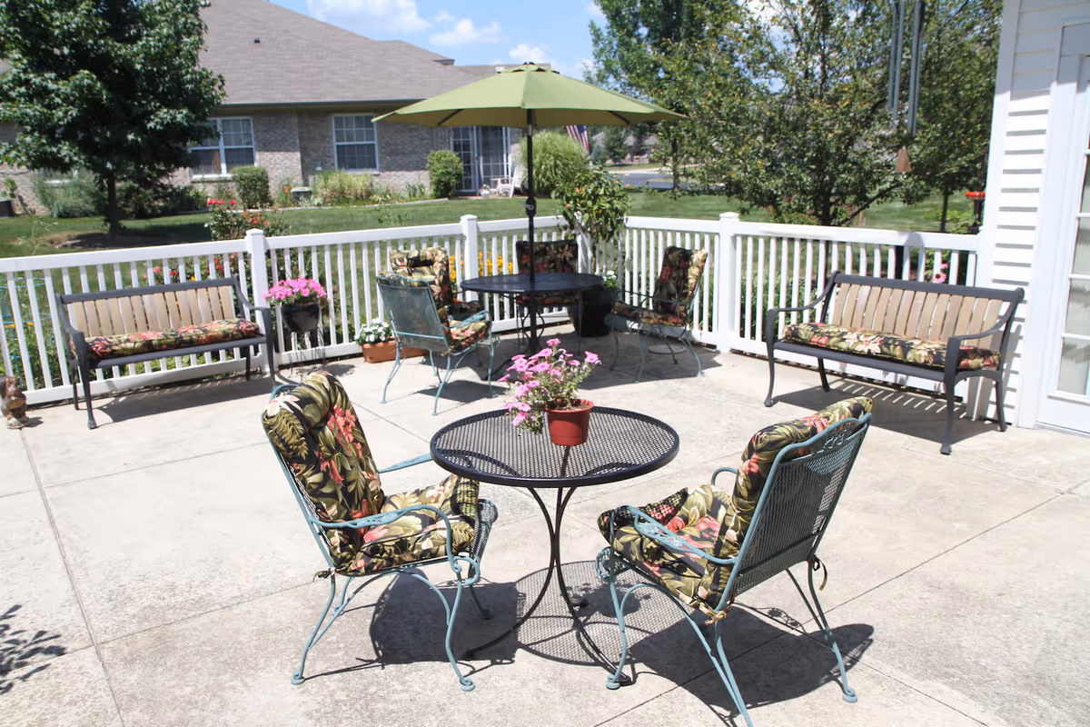 Outdoor patio area with metal chairs and benches featuring floral cushions, a round table with a potted plant, and a green umbrella. The patio is surrounded by a white fence and greenery, with a building and lawn visible in the background.