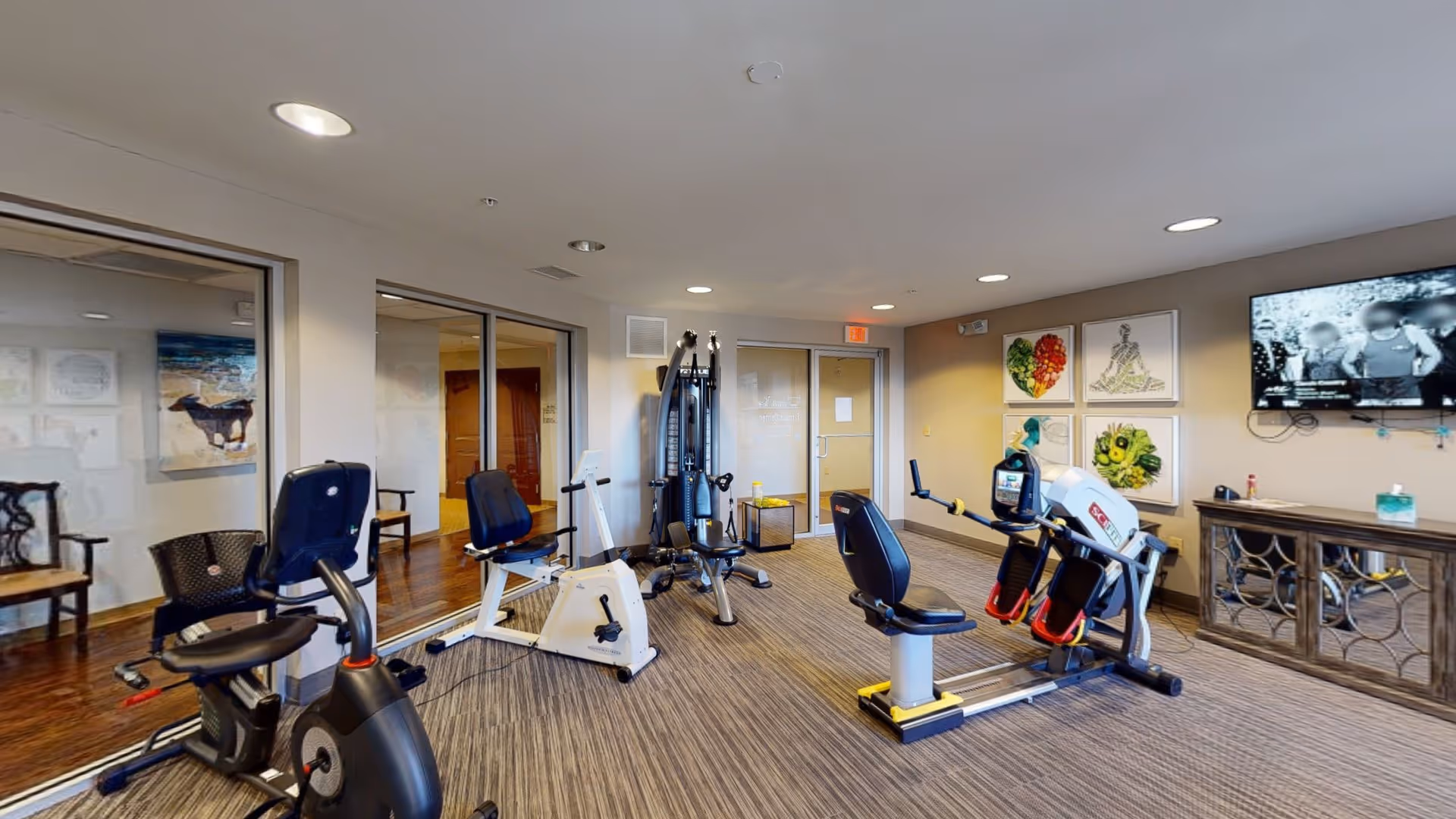 A fitness room in a senior living facility with exercise equipment including stationary bikes, a multi-function weight machine, and a rowing machine. The room has carpeted flooring, recessed ceiling lights, and framed artwork on the walls. A flat-screen TV is mounted on the wall above a wooden cabinet.