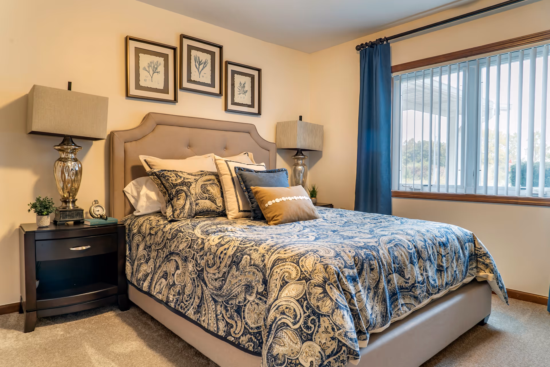 A neatly made bed with a beige upholstered headboard and blue paisley-patterned bedding in a bedroom. There are multiple pillows arranged on the bed. On each side of the bed are dark wooden nightstands with silver lamps and small decorative plants. Above the bed are three framed botanical prints. A large window with vertical blinds and blue curtains is on the right side of the room.