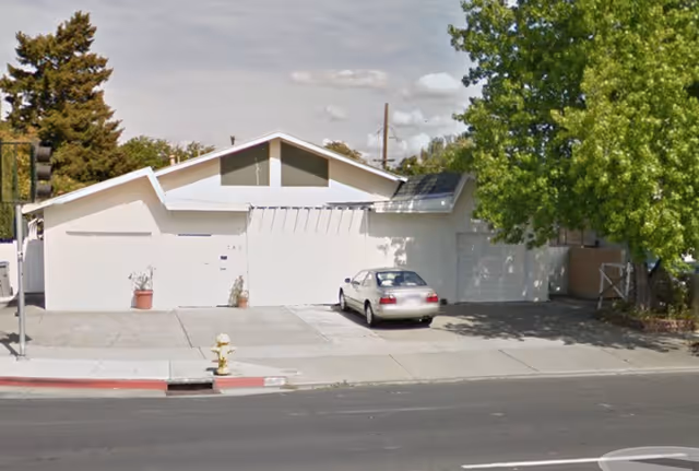 Exterior view of a single-story white building with a slanted roof, a parked silver car in the driveway, a large green tree on the right side, and a fire hydrant near the curb in front.
