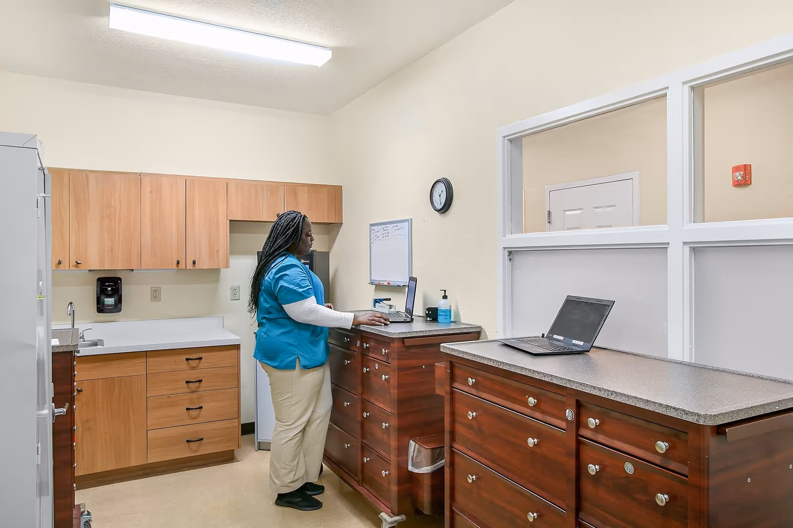 A healthcare worker in blue scrubs and beige pants stands in a small office or workroom with wooden cabinets and countertops. She is using a laptop placed on a wooden cabinet. The room has a clock on the wall, a whiteboard, and a hand sanitizer dispenser. There are two laptops on the cabinets and a sink with a soap dispenser on the left side.