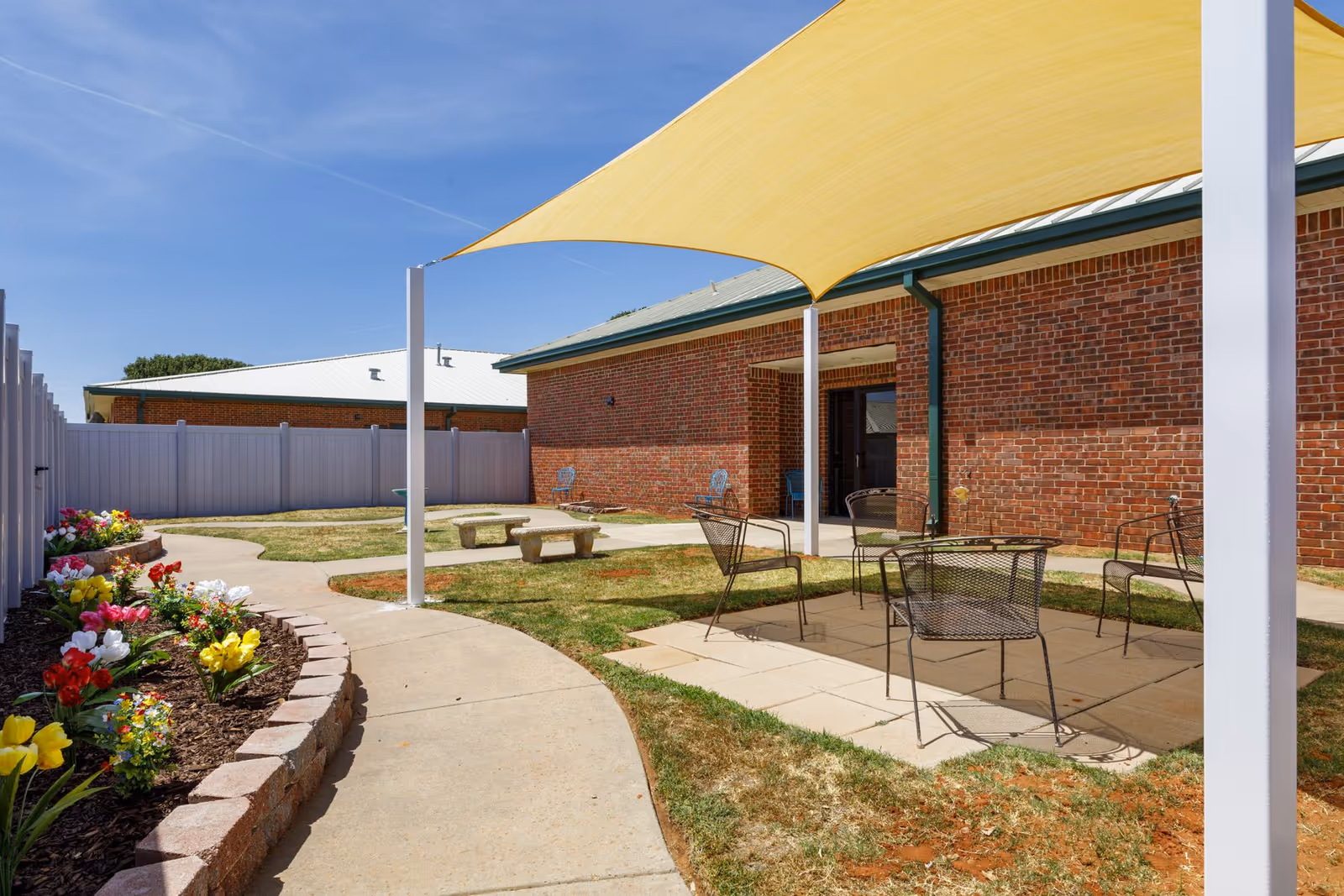 Outdoor patio area at Homestead Senior Living of Elk City featuring a yellow shade sail, metal chairs, a concrete bench, a curved walkway, and a flower bed with colorful flowers along a white fence. The building has a brick exterior with a green roof.