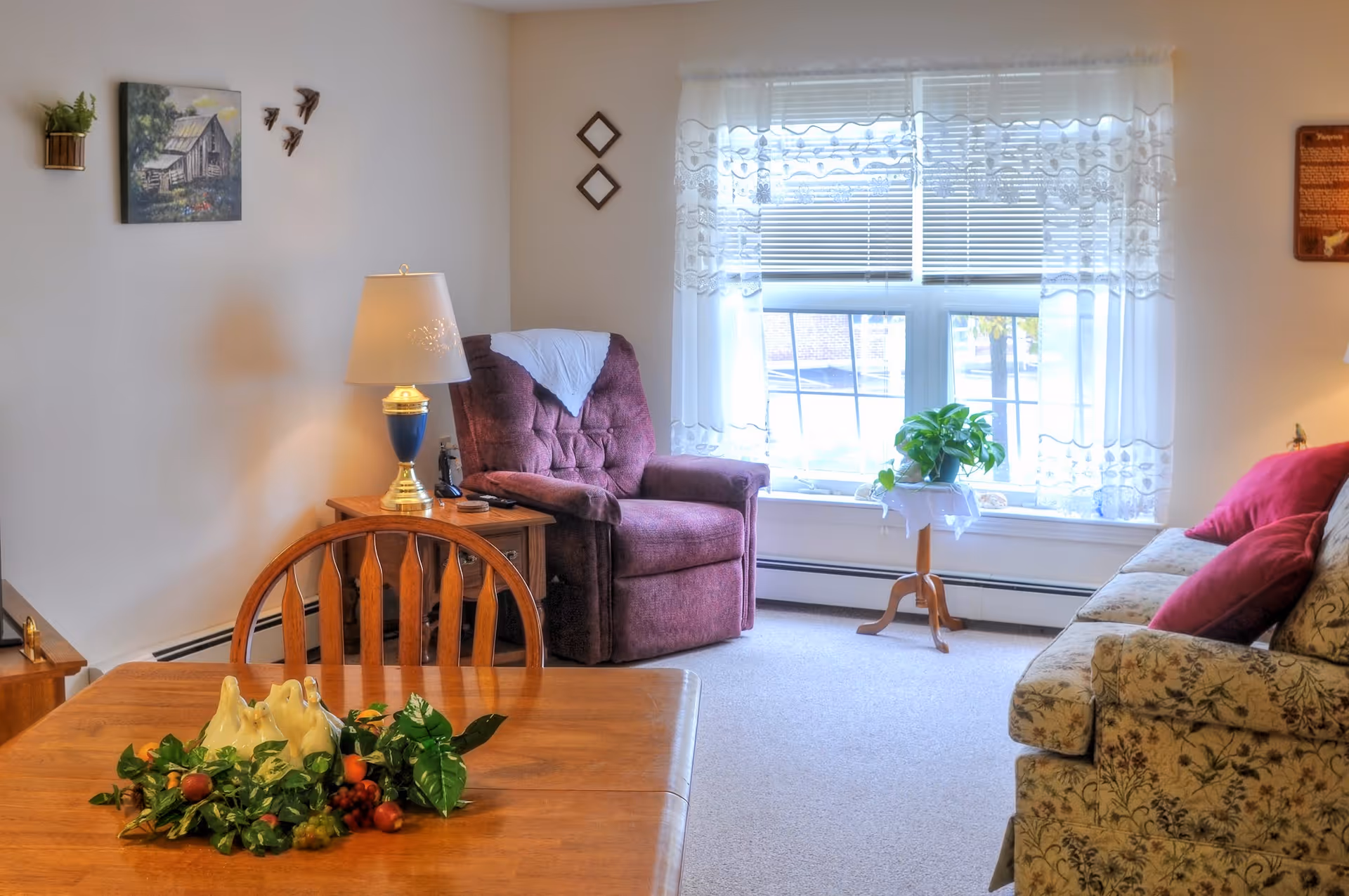 A cozy living room area with a purple armchair next to a wooden side table with a lamp, a floral patterned sofa with red cushions, a wooden dining table with a decorative centerpiece of ceramic birds and greenery, and a window with sheer white curtains letting in natural light. The walls are decorated with small framed pictures and wall-mounted plants.