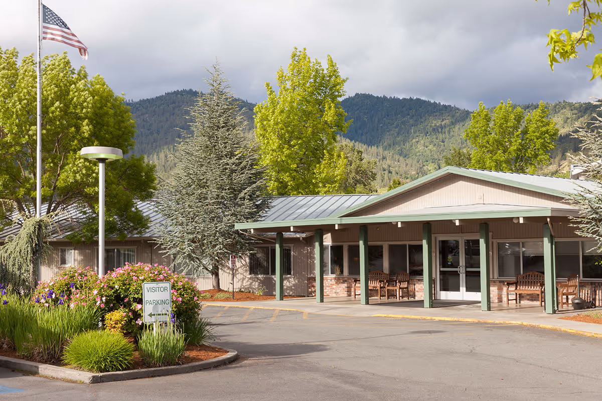 Front exterior of a single-story health and rehabilitation center with a covered entrance, benches, landscaped flower beds, an American flag, and tree-covered hills in the background.