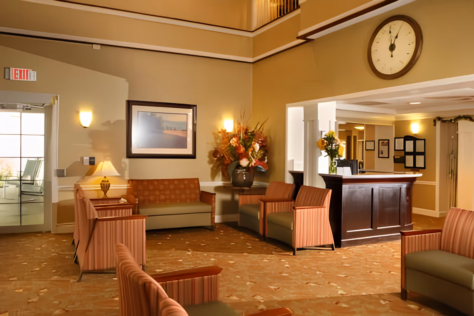 Lobby seating area with upholstered chairs and sofas, a reception desk, wall clock, and a floral arrangement.