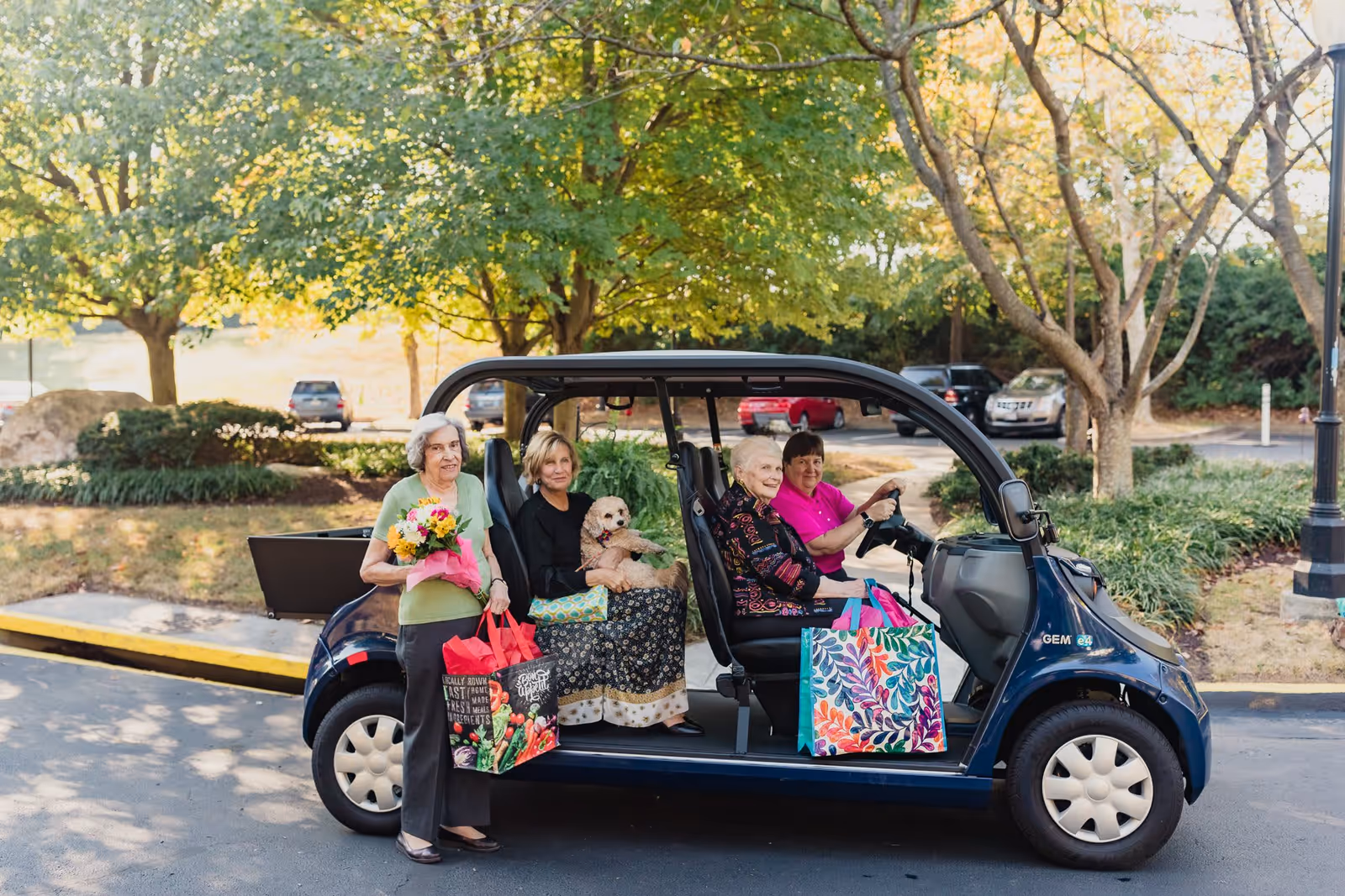 Four elderly women enjoying a ride in a small open electric vehicle in a senior living community parking area. One woman is standing outside holding a bouquet of flowers and a shopping bag, another is seated inside holding a small dog, and two others are seated in the vehicle with colorful shopping bags. Trees and parked cars are visible in the background.