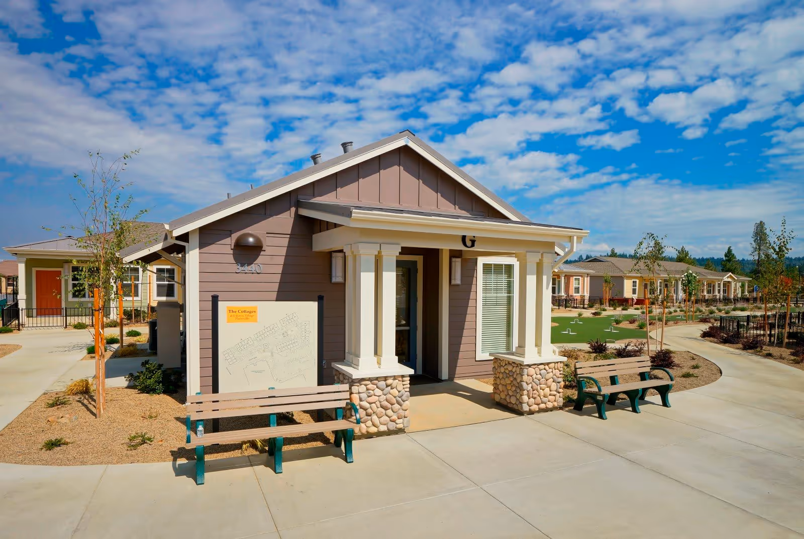 Exterior view of a small building with a covered entrance supported by columns with stone bases, two benches on either side of the entrance, a pathway, and landscaped areas with young trees and shrubs under a partly cloudy blue sky.