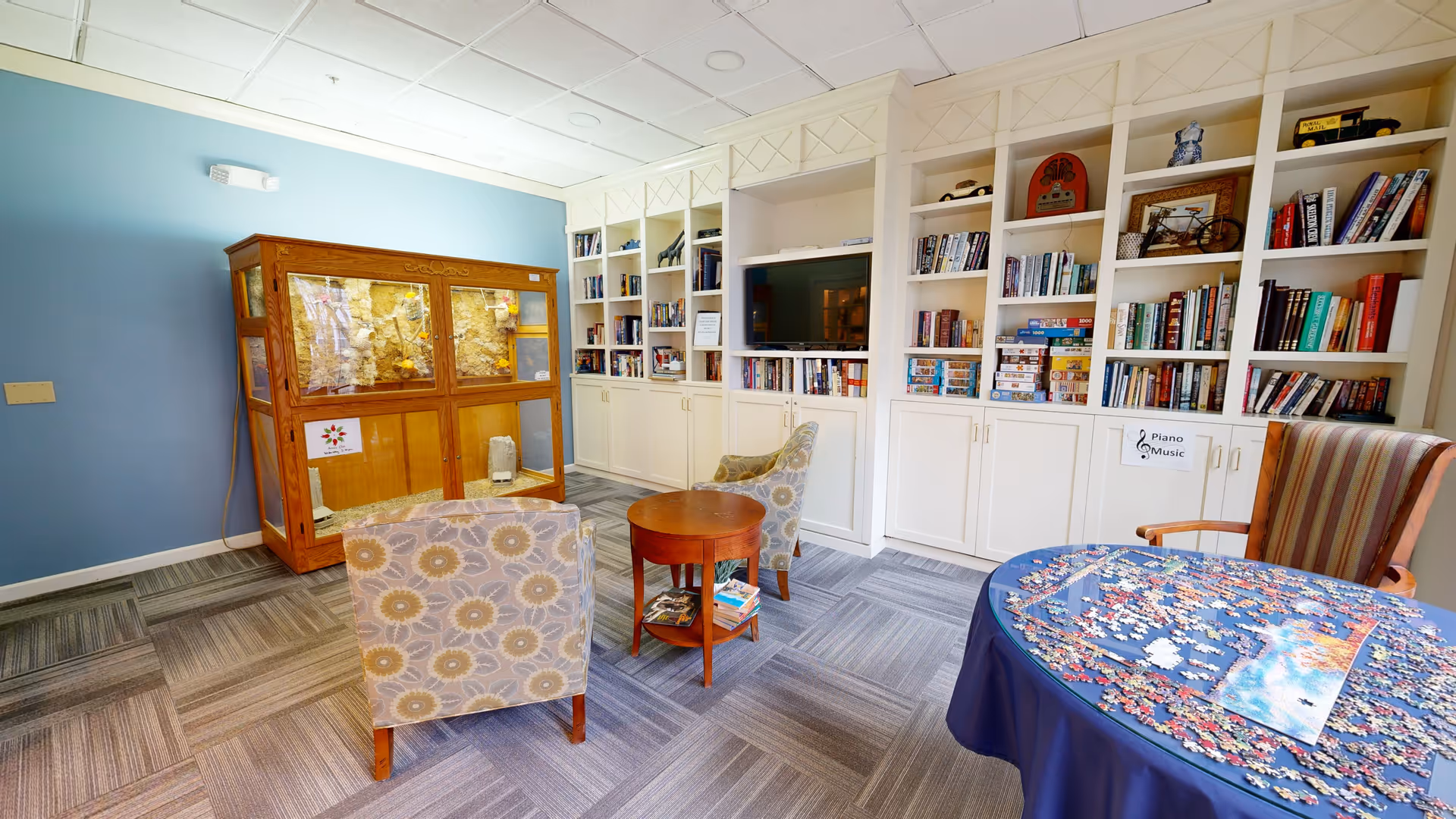 Bright common lounge with built-in bookshelves, armchairs, a round table with a jigsaw puzzle, and a wooden display cabinet against a blue wall.