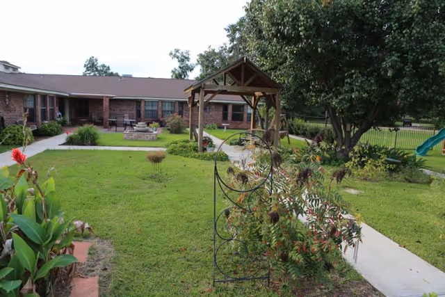 Grassy courtyard featuring a wooden gazebo, walkways, landscaping, and a surrounding single-story brick nursing facility.