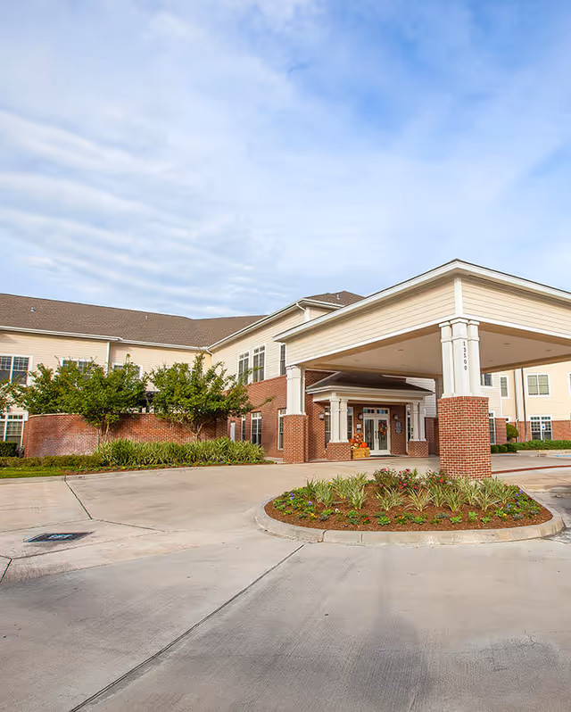 Exterior view of a senior living facility with a covered entrance supported by brick and white columns, landscaped flower beds, and a clear blue sky with some clouds.