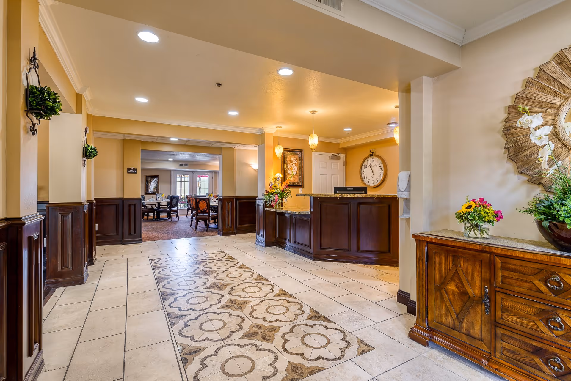 Interior view of a senior living facility lobby area with a decorative tiled floor, a wooden reception desk with flowers, a large wall clock, and a wooden sideboard with floral arrangements. In the background, there is a dining area with tables and chairs and large windows letting in natural light.