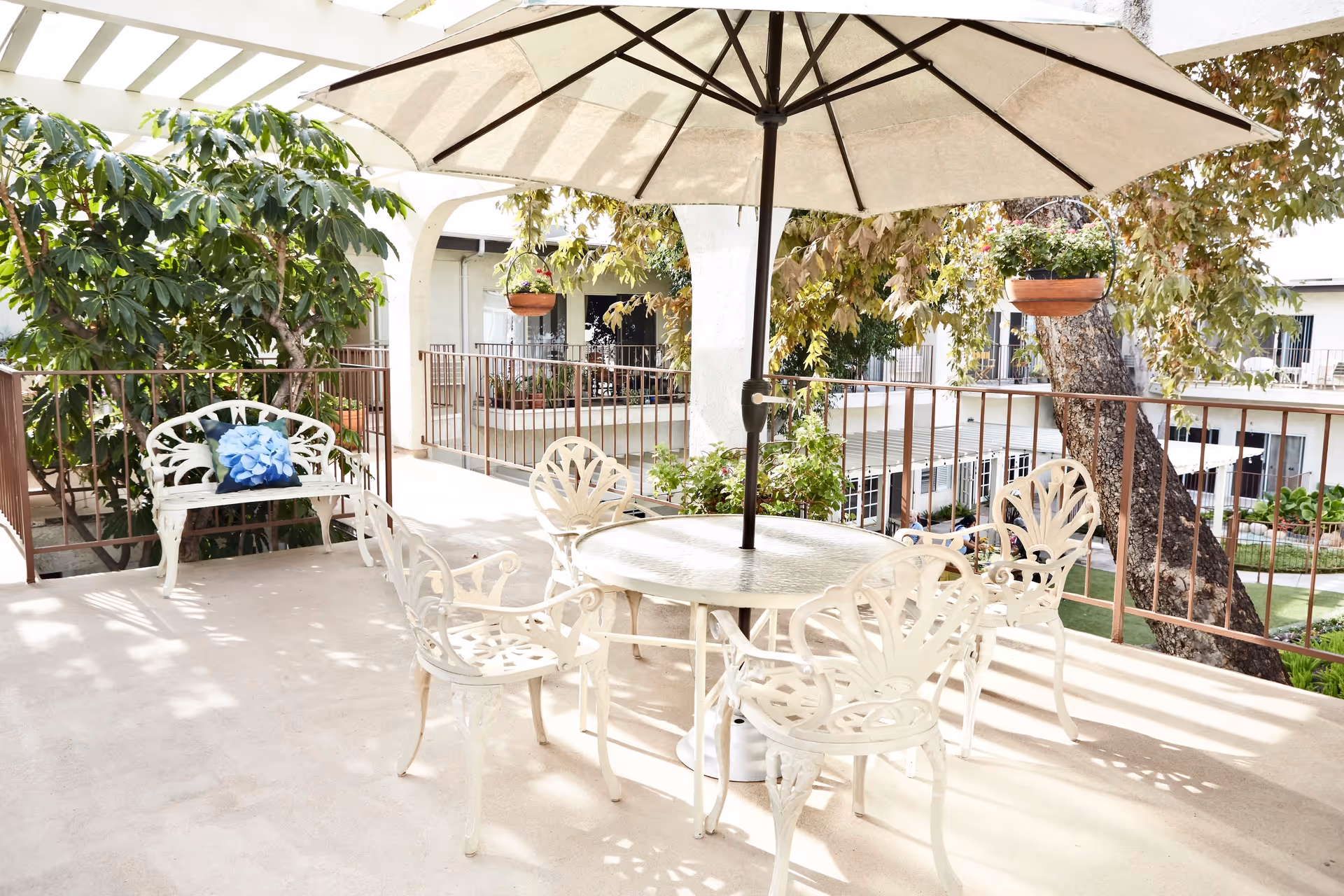 Outdoor patio area with a round glass table and four white metal chairs under a large beige umbrella. There is a white metal bench with a blue floral cushion on the left side, surrounded by green plants and trees. The patio is elevated with a metal railing and overlooks a courtyard with more greenery and building windows.