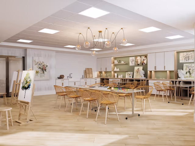 A bright and spacious art room with wooden flooring, multiple wicker chairs around tables, easels with paintings, and shelves displaying sculptures and art supplies. The room has modern lighting fixtures and a clean, organized appearance.