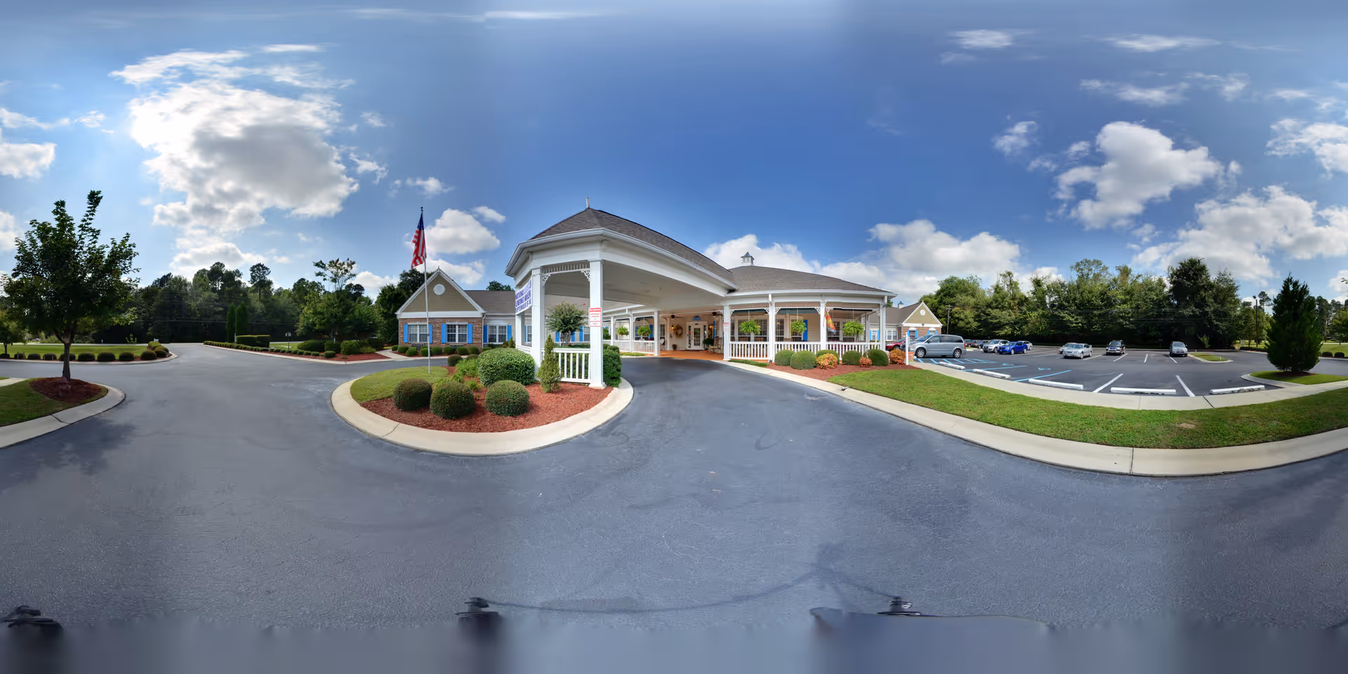 Exterior view of The Legacy of Orangeburg senior living facility showing the main entrance with a covered drop-off area, landscaped bushes, trees, and an American flag. The parking lot is visible with several parked cars under a partly cloudy blue sky.