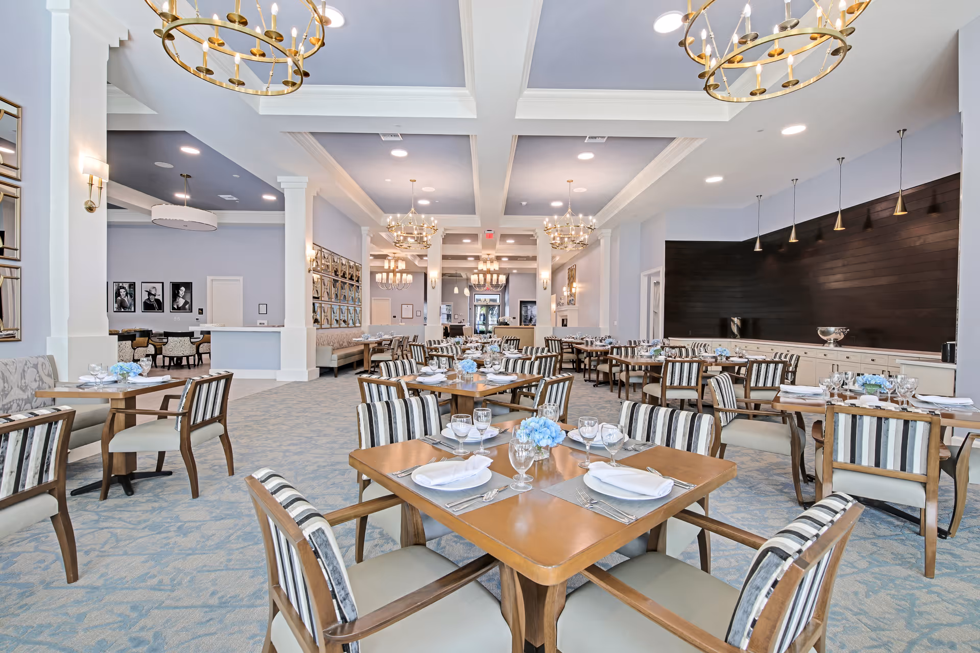A spacious and elegant dining room in a senior living facility with multiple wooden tables set with white napkins, glassware, and blue floral centerpieces. The room features striped cushioned chairs, light blue carpeting, white walls, and large gold chandeliers hanging from a coffered ceiling. There are framed pictures on the walls and a dark wood accent wall with pendant lights on the right side.