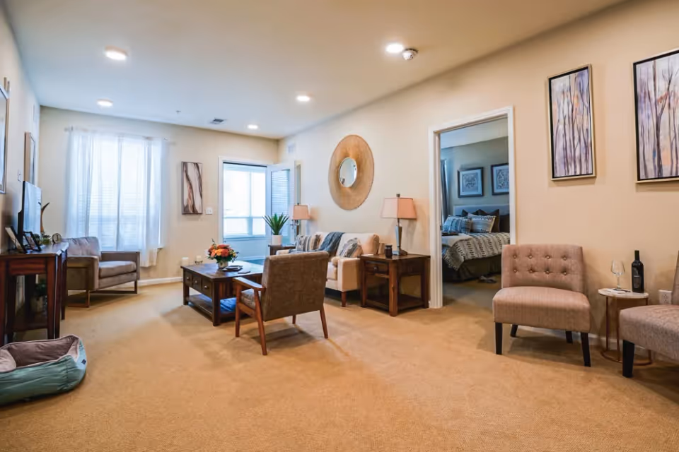 A cozy living room with beige carpet and walls, featuring a sofa with cushions, two armchairs, a wooden coffee table with a flower arrangement, a TV on a wooden stand, and a doorway leading to a bedroom with a neatly made bed. The room is decorated with framed artwork, a round mirror, and table lamps, with natural light coming through a window with sheer curtains.