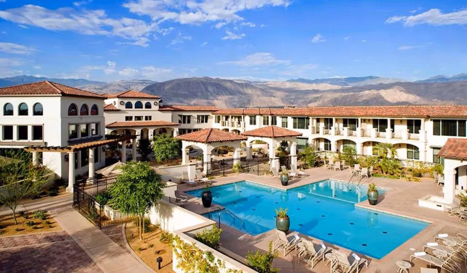 Mediterranean-style senior living complex courtyard with a central swimming pool, lounge chairs, and two-story buildings set against a mountain backdrop.