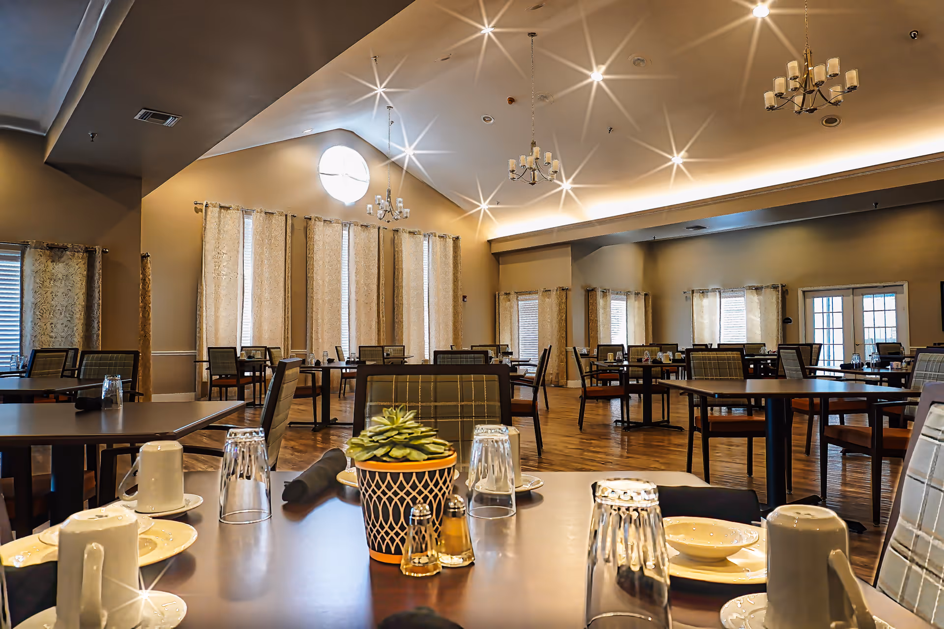 A spacious dining room with multiple tables and chairs arranged neatly. Each table is set with plates, cups, upside-down glasses, and black napkins. A small potted plant and salt and pepper shakers are on the table in the foreground. The room features high ceilings with chandeliers and large windows covered with patterned curtains, allowing natural light to brighten the space.