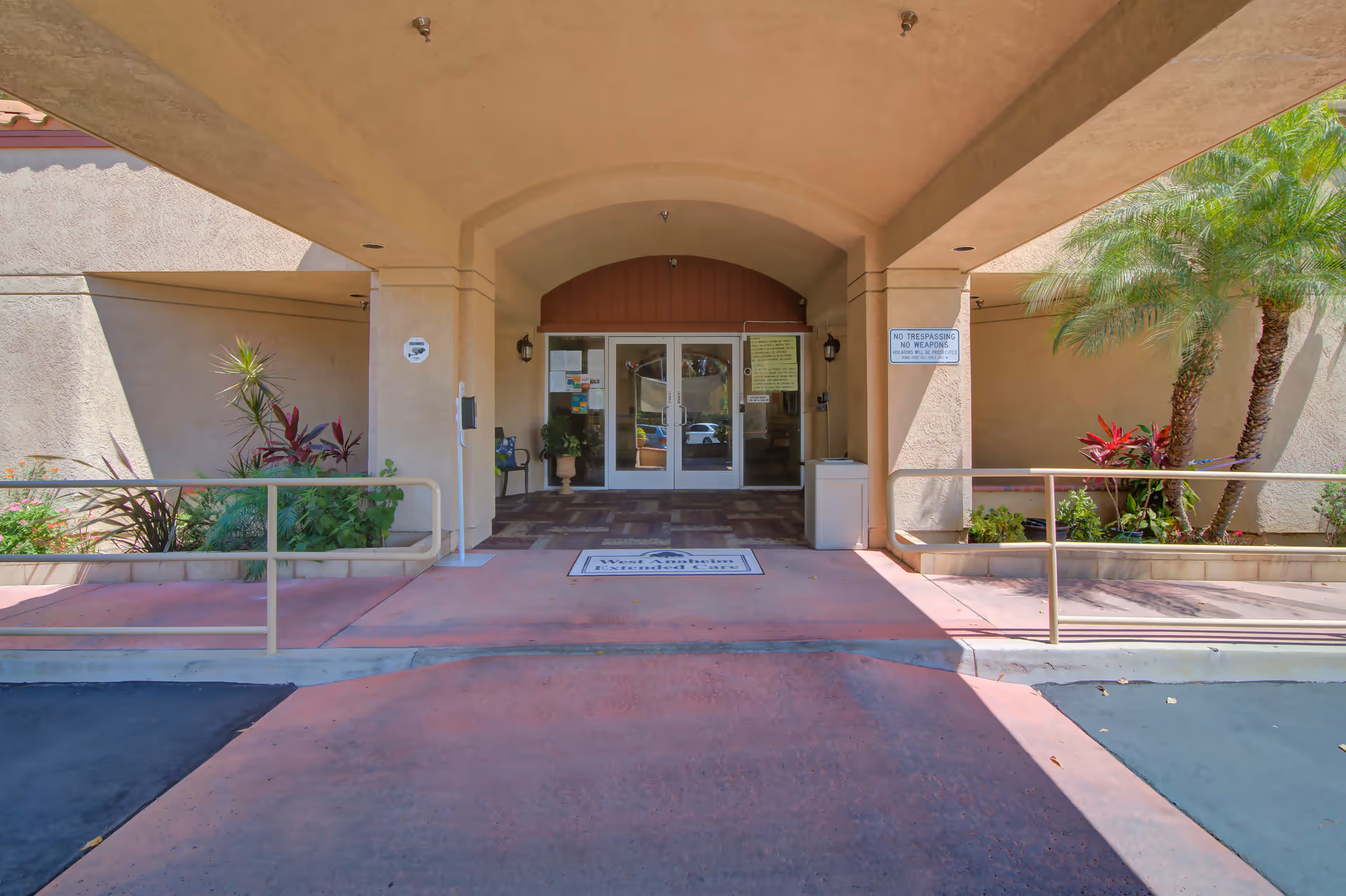 Entrance to a building with a covered walkway, glass double doors, potted plants, and a ramp with handrails on both sides. There are signs on the walls near the doors and landscaping with palm trees and other plants on either side of the entrance.