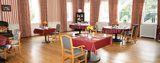 A dining room with several tables covered in red tablecloths, each set with plates, glasses, and floral centerpieces. The room has large windows with striped curtains, wooden flooring, and chairs with cushioned seats around the tables.