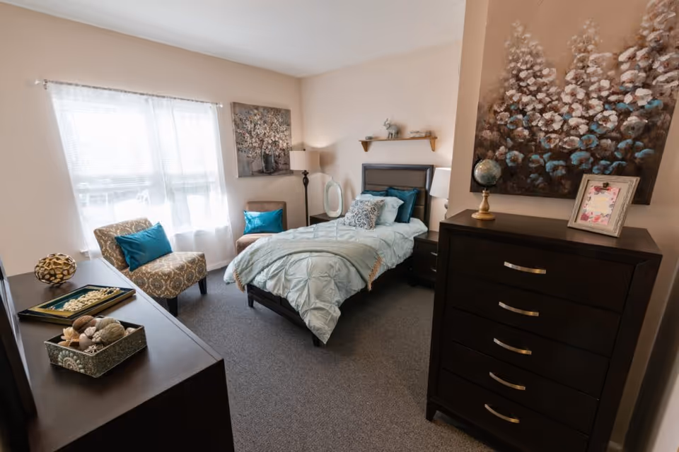 A cozy bedroom with a single bed made with light blue bedding and decorative pillows. The room features a window with sheer white curtains, two patterned chairs with blue accent pillows, a dark wooden dresser with a globe and framed picture on top, and a matching nightstand with a lamp. The walls are decorated with two large paintings.
