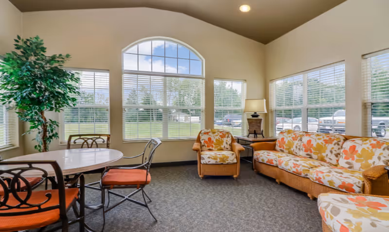 A bright and cozy living room area with large windows letting in natural light. The room features a floral-patterned sofa and matching armchair, a round table with four chairs that have orange cushions, a tall green potted plant, and a side table with a lamp. Outside the windows, a grassy area and parked vehicles are visible.