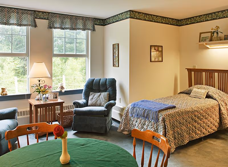A cozy room with a single bed covered in a patterned quilt and a blue throw blanket. Next to the bed is a wooden headboard with a small shelf holding framed photos and a plant. There is a comfortable blue armchair with a decorative pillow beside a wooden side table with a lamp, flowers, and framed photos. Two large windows with floral valances let in natural light, and a round table with a green tablecloth and wooden chairs is in the foreground.